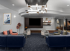 Modern hotel lobby with dark blue sofas, armchairs, and a central TV. Contemporary light fixture and decorative shelves enhance the space.