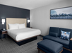 Modern hotel room with a large bed, two bedside tables with lamps, a blue sofa, an ottoman, and a framed picture on the wall.