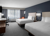 Hotel room with two large beds, a work desk with chair, an orange armchair, a wall-mounted TV, and a window with sheer curtains.