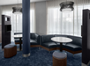 Modern lounge area with dark blue cushioned seating, round tables, tall partitions, and hanging light fixtures. Large windows with sheer curtains provide natural light.