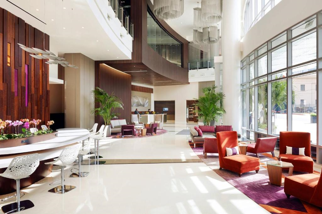 Modern hotel lobby with large windows, red and white seating, and indoor plants. A reception desk is visible in the background. Natural light fills the space.