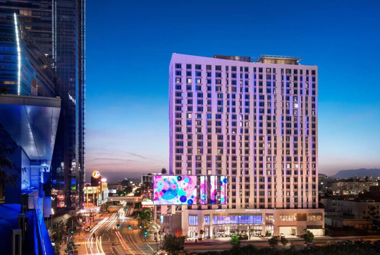 Urban street scene at dusk featuring a modern, tall building illuminated with colorful lights.