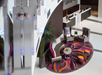 Modern hotel lobby with a circular seating area, two armchairs, a curved sofa, a round table, and colorful carpet. Reception desk in the background.