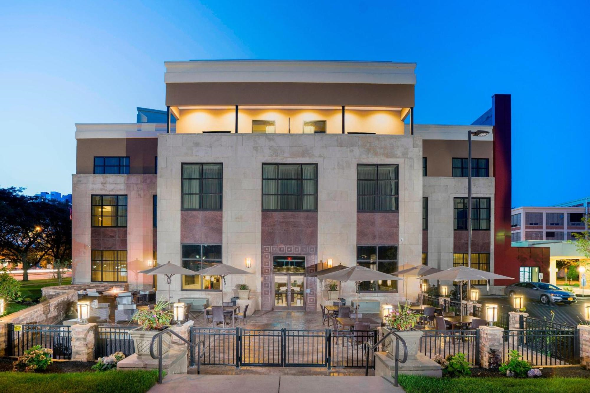 A modern three-story building with large windows and an outdoor patio area featuring tables, chairs, and umbrellas, photographed at dusk.