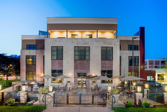 A modern three-story building with large windows and an outdoor patio area featuring tables, chairs, and umbrellas, photographed at dusk.