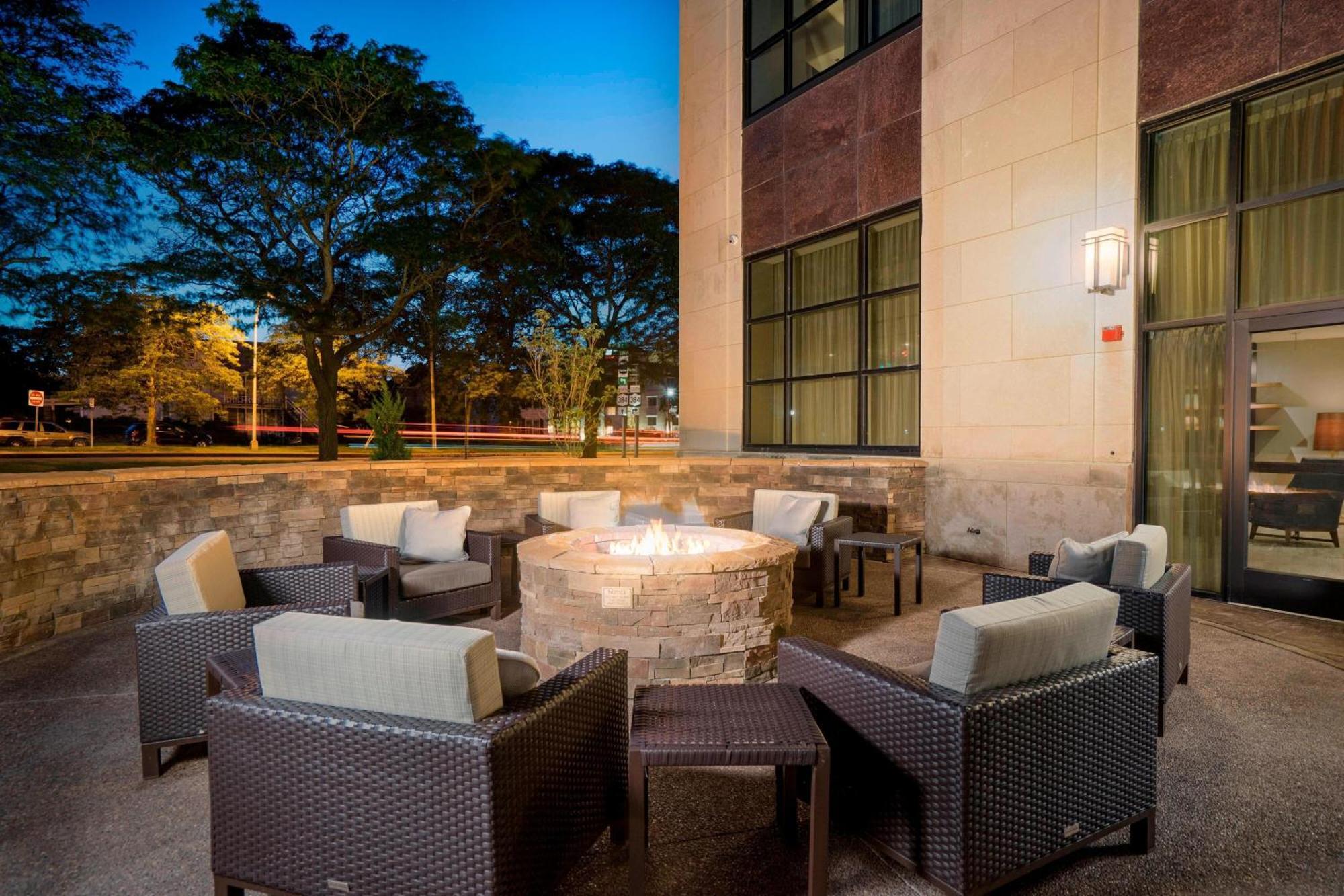 Outdoor patio area with wicker chairs arranged in a circle around a lit stone fire pit, next to a building with large windows, at dusk.