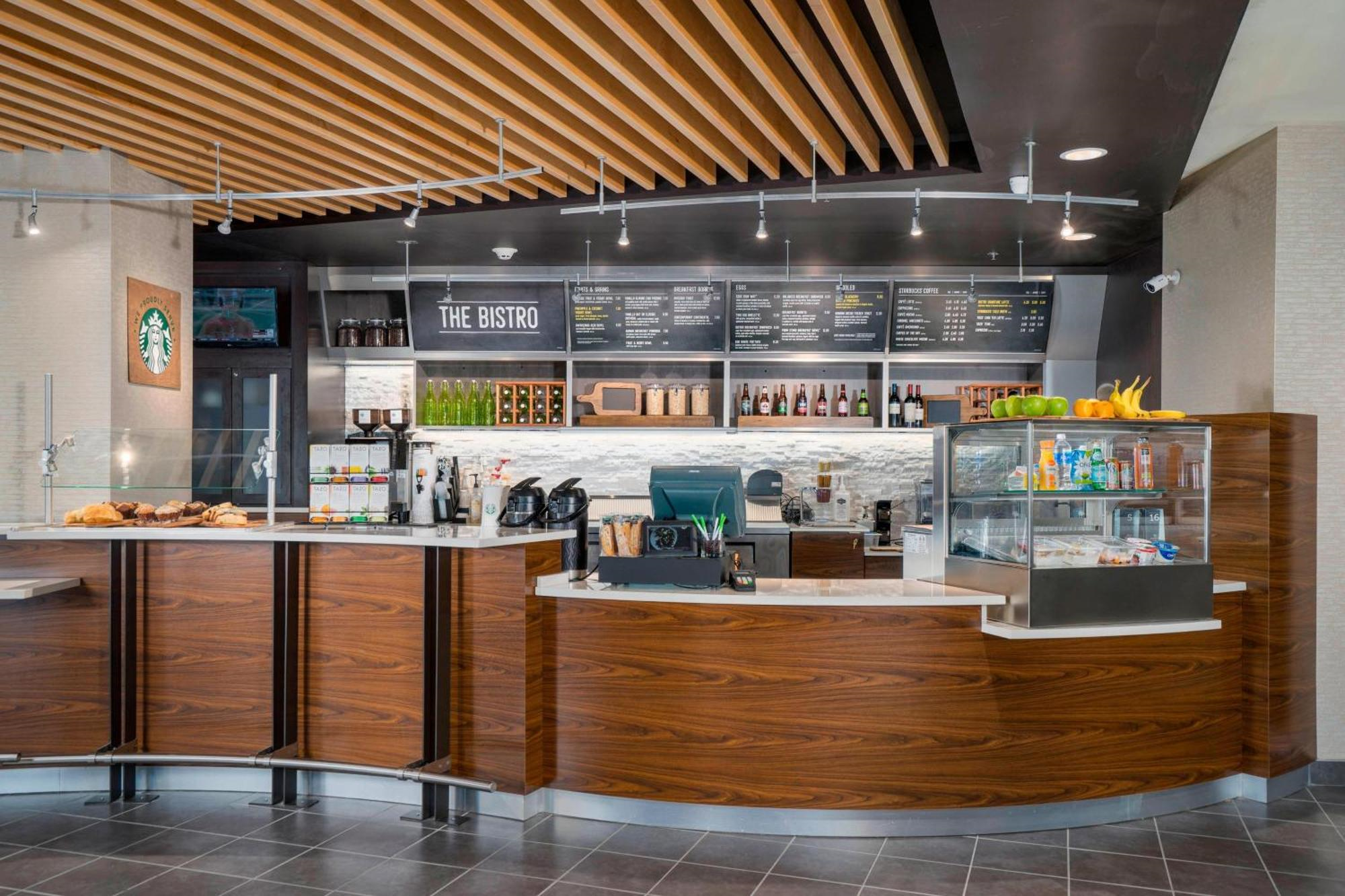 Modern café counter with wood accents, display case with snacks, coffee machines, and menu boards overhead; Starbucks logo visible on the left.