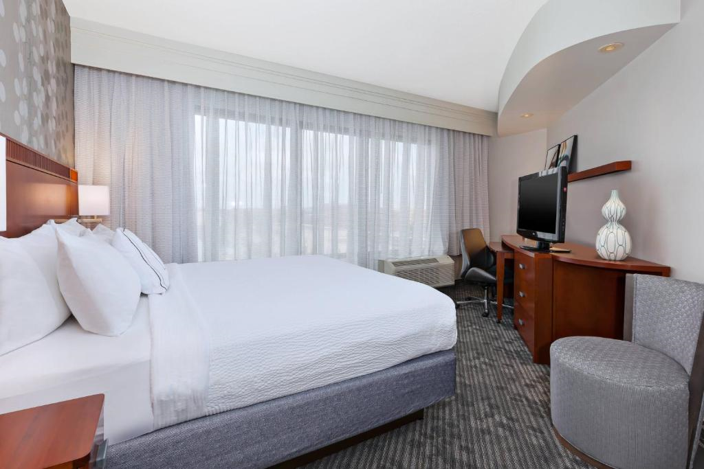 A hotel room features a neatly made bed, light gray curtains, a flat-screen TV on a wooden stand, a desk with a chair, and decorative items. The carpet is patterned in subtle gray tones.