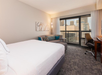 Hotel room with a large bed, a sofa, a desk with a chair, and a window showing a city skyline. Walls are light-colored, and the floor is carpeted.