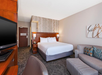 Hotel room with a king bed, two bedside tables with lamps, a wall-mounted TV, a sofa, and armchair. Walls are light-colored with patterned wallpaper behind the bed.
