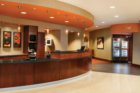 Hotel lobby with a curved wooden reception desk, wall art, and a small market area featuring refrigerated drinks. The ceiling and lighting are modern with orange accents.