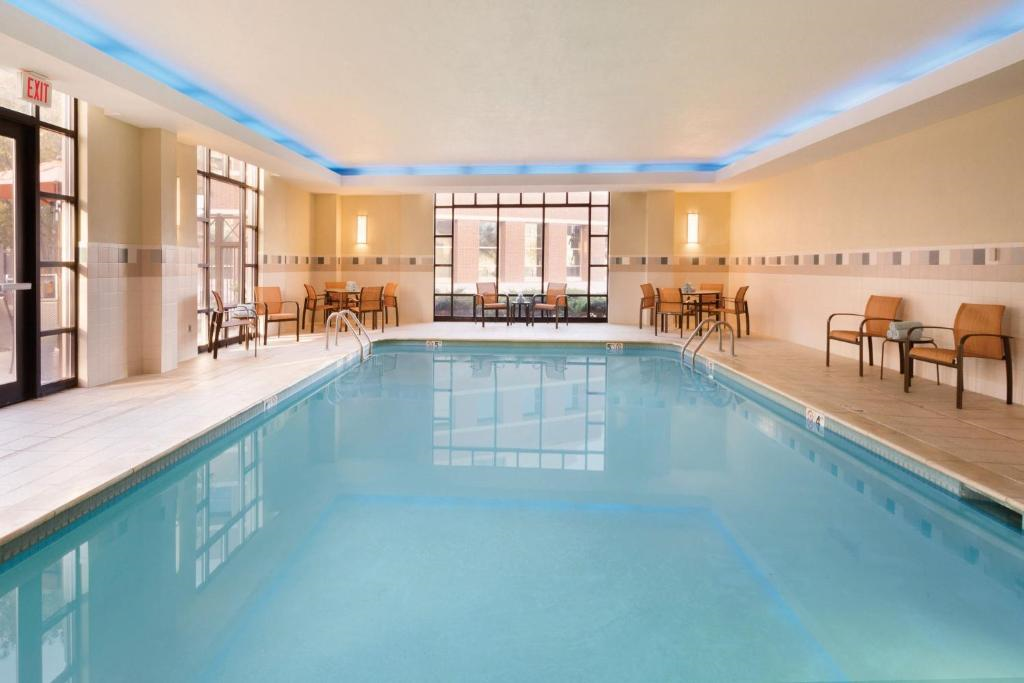 Indoor swimming pool with clear water, surrounded by chairs and tables along the walls, under soft lighting and a large window at the far end.