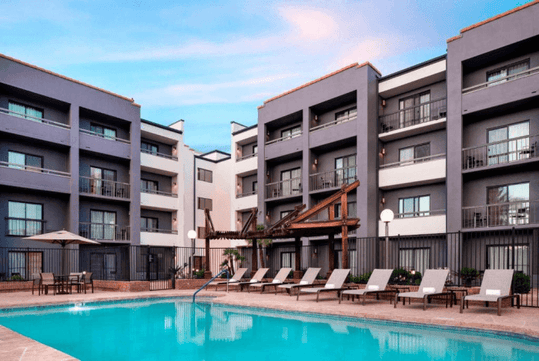Four-story hotel with balconies surrounding an outdoor pool. Lounge chairs and a patio area with umbrellas are visible.