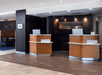 Hotel lobby with two wooden reception desks, each with a computer monitor. The background features a dark wall with decorative elements. The floor is a mix of wood and tile.