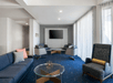 A modern lounge with blue carpet, gray sofas, armchairs, a wall-mounted TV, and a glass coffee table with a decorative piece.