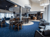 Modern lobby with dark blue carpet, wooden chairs, and tables. Circular ceiling design and hanging lights. Reception desk and seating area in the background.