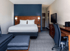 Modern hotel room with a bed, desk, TV, chair, and sofa. The wall behind the bed is dark blue, and there's a hallway leading to the exit. A coffee maker sits on the desk.
