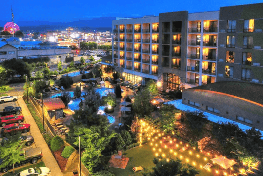 Aerial view of a hotel with an illuminated courtyard, swimming pool, and surrounding greenery at dusk.