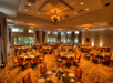 A banquet hall with multiple round tables set for an event. Elegant chandeliers hang from the ceiling, and large windows line the walls, providing natural light.