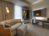 Modern hotel room with a sofa, ottoman, floor lamp, desk, office chair, wall-mounted TV, and a window with curtains.