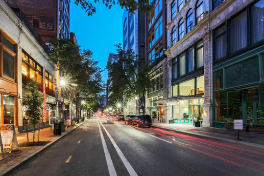 Street view at dusk with lit buildings, parked cars, and blurred taillights. Trees line the sidewalk, and the sky is twilight blue.
