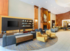 Spacious hotel lobby with modern furniture, including yellow and gray seating, a wall-mounted TV, shelves with decorative items, and a fireplace.