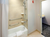 Accessible bathroom with a bathtub featuring grab bars, a showerhead above, and a white towel on the tub edge. An open door leads to a carpeted area with a countertop.