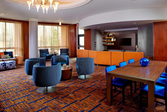 Modern hotel lobby with blue and orange decor, featuring circular chairs, a wooden table with blue chairs, a TV, and ambient lighting.