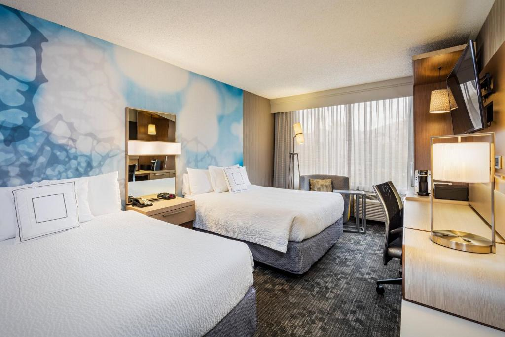 Hotel room with two double beds, a blue accent wall, a desk, a TV, a floor lamp, and a large window with curtains.