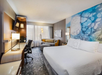 Modern hotel room with a large bed, desk, chair, sofa, and abstract wall art. Room features a mounted TV, floor lamp, and a window with sheer curtains.