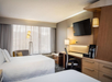 Hotel room with two beds, a work desk with a chair, wall-mounted TV, floor lamp, and coffee maker, featuring neutral tones and large window with sheer curtains.