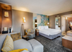 Modern hotel room with a king-sized bed, sofa, and a work desk. The decor features neutral tones with accent lighting and abstract wall art.