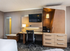 Hotel room workspace with a modern wooden desk, a black office chair, a wall-mounted TV, a desk lamp, and a coffee maker. A tall mirror is in the corner.