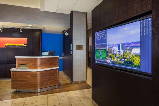 Hotel lobby with a wooden reception desk and a wall-mounted TV displaying local information and images.