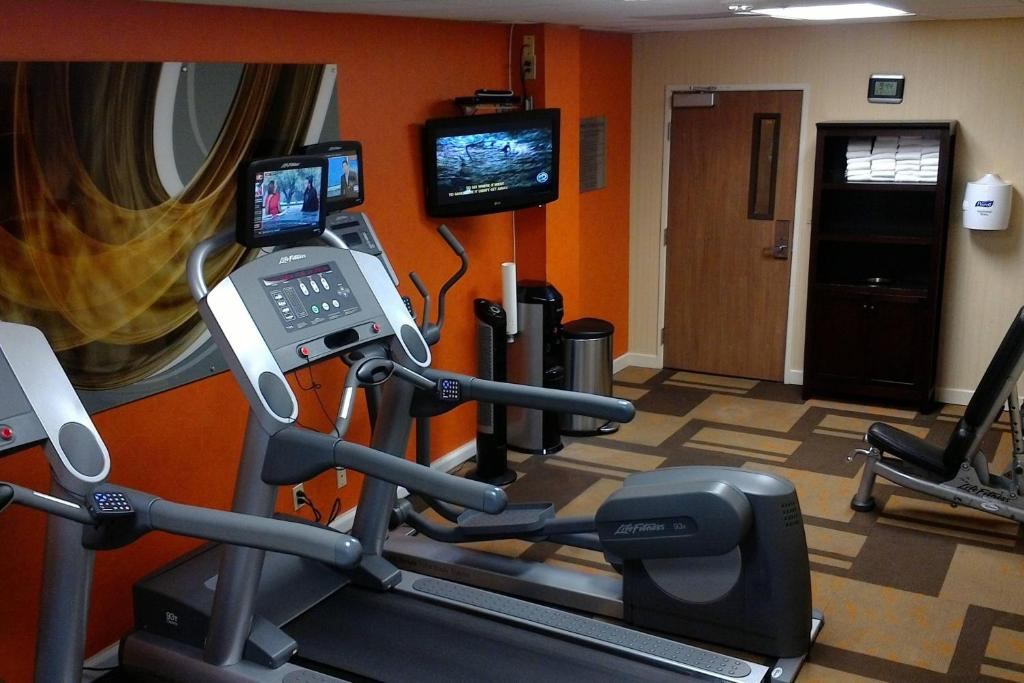 Small gym room with two treadmills, a stationary bike, two wall-mounted TVs, a shelf with towels, and an abstract art piece on an orange wall.