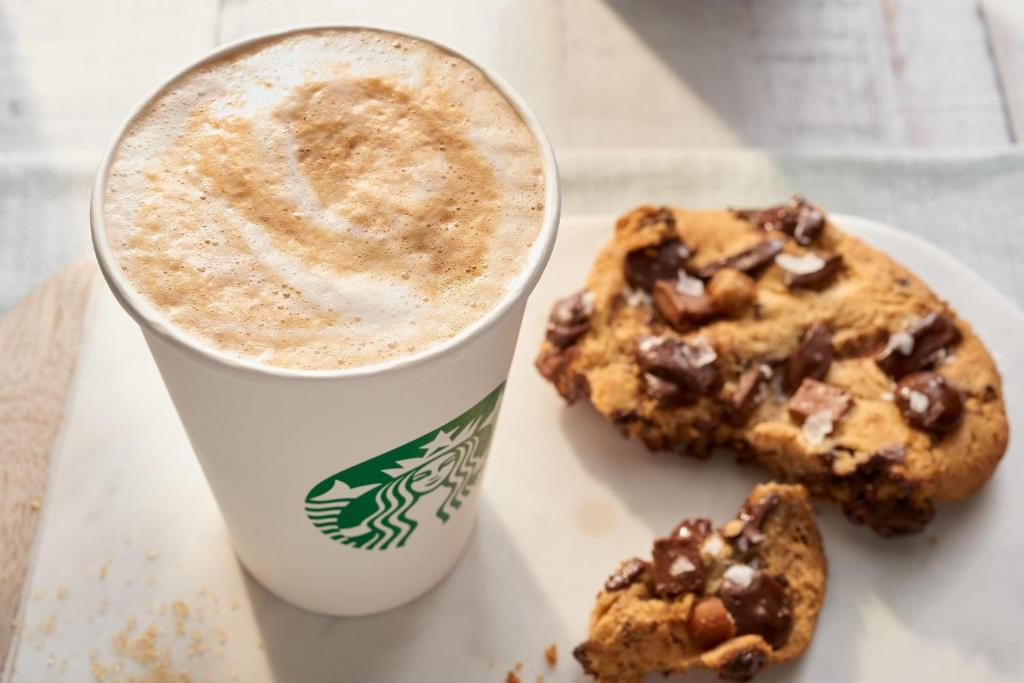 A cup of frothy coffee next to a chocolate chip cookie, some pieces broken off, on a white surface.