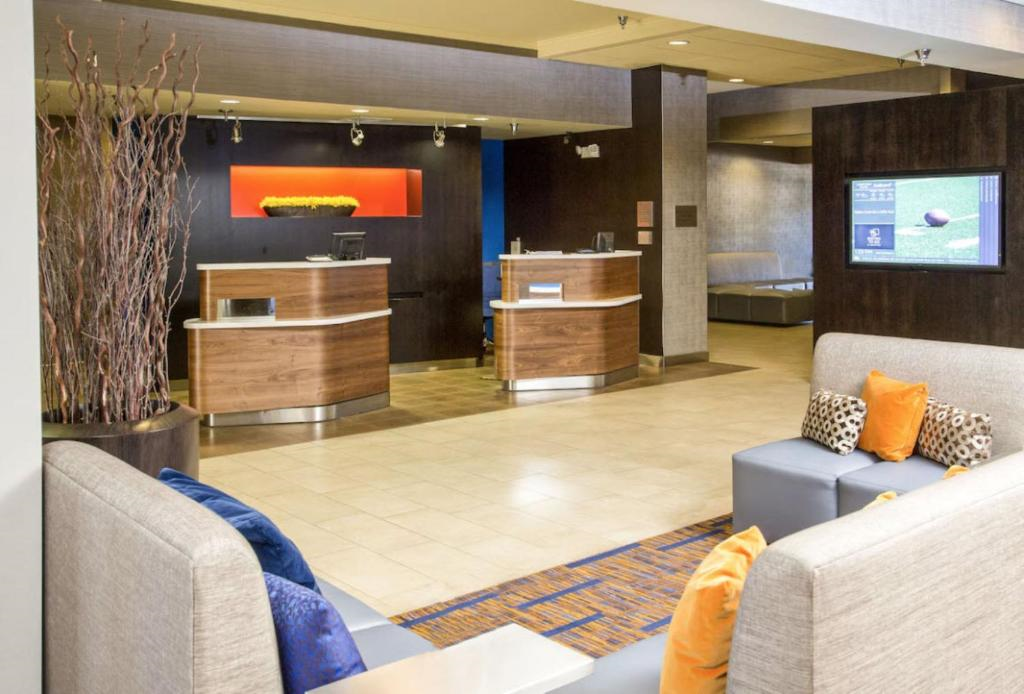 Modern hotel lobby with light tile floors, two curved wooden reception desks, decorative branches, and seating area with gray couches and orange and patterned pillows.