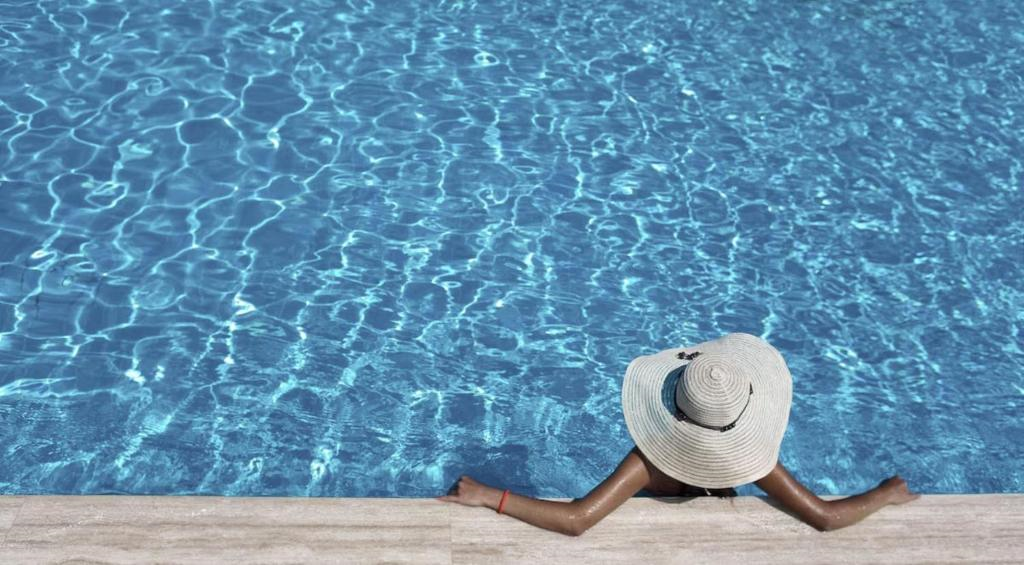 Person wearing a wide-brimmed hat sits at the edge of a swimming pool with arms resting on the poolside, facing the water.