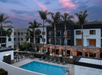 A modern hotel with palm trees, a pool, and lounge chairs. Warm string lights illuminate the area. The sky is partly cloudy with pink and purple hues at dusk.
