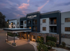 Exterior view of a Courtyard by Marriott hotel with a modern facade and entrance, surrounded by trees and plants, under a dusk sky.