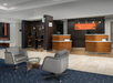 Modern hotel lobby with two gray chairs, a glass table, and a reception desk. There are computers for guest use labeled "Boarding Pass.