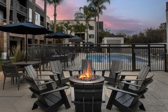 Outdoor seating area with black chairs around a fire pit, near a pool. Tall palm trees and a building are in the background under a pink sunset sky.