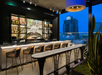 Rooftop bar at dusk with a long counter, high chairs, colorful bottles on shelves, and cityscape view through large windows.