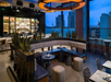 Rooftop bar with modern decor, featuring a curved seating area, tables set with plates and cutlery, a bar with a lit display of bottles, and city skyline views through large windows.