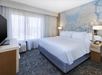 A modern hotel room with a large bed, two bedside tables with lamps, a flat-screen TV, and a window with sheer curtains. The wall features abstract artwork.