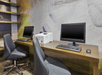 Office workspace with two computers, a printer, and chairs at a wooden desk. Shelving unit with decor visible on the left.