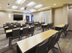 An empty conference room with rows of tables and chairs facing a large screen and podium under a white ceiling with recessed lighting.