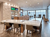 Modern hotel lobby with a spacious seating area, bar, and wall-mounted TVs. Neutral tones, comfortable chairs, and large windows create a welcoming atmosphere.