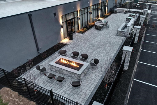 Outdoor patio with a fire pit, surrounded by eight chairs. Adjacent is a modern building with large windows.