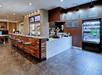 Modern cafe interior with a wooden counter, bar stools, digital menu boards, and a beverage cooler against a dark wood-paneled wall.